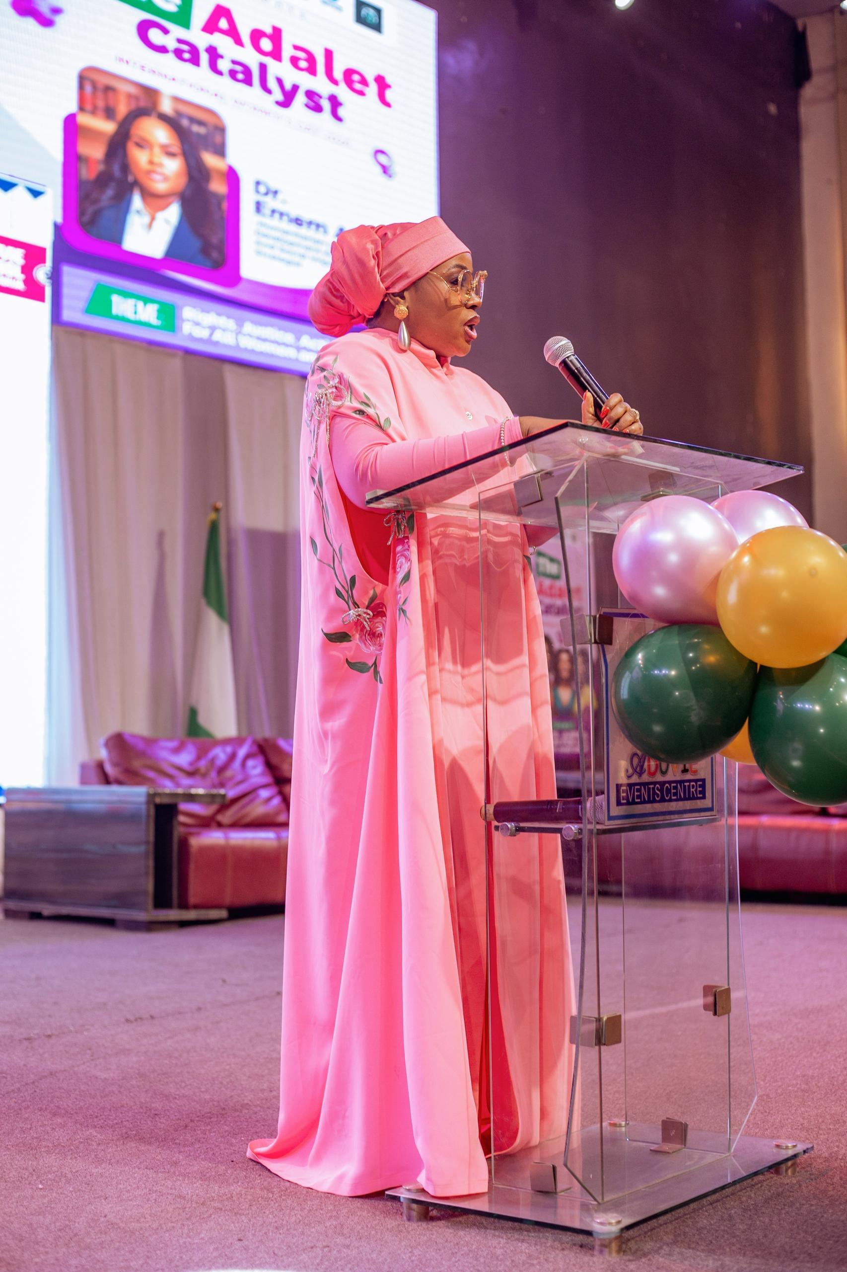 Dr Emem Alban speaking into a microphone at a lectern, wearing a pink outfit and matching headwrap